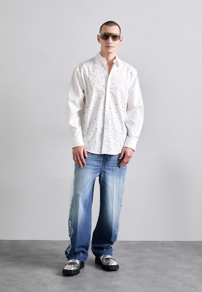 Weißes Hemd mit knöpfen und bunten Punkte-Mustern, kombiniert mit hellen blauen Jeans mit dekorativen Elementen. Das Model trägt verspiegelte Sonnenbrillen und metallische Schuhe.
