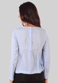 Blusa de manga larga color azul claro, con una textura suave, escote redondeado y cierre de botones en la parte de atrás. Presenta un corte relajado y dobladillo.