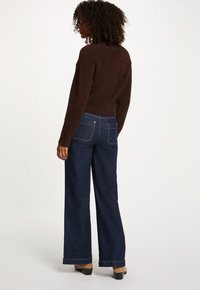 Brauner, flauschiger Pullover und weite, dunkelblaue Jeans. Die Jeans haben zwei Gesäßtaschen und kontrastierenden Nähten. Stiefel sind darunter sichtbar.
