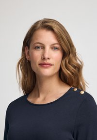 Mörkblå topp i mjukt tyg med rund halsringning och två gyllene knappar på axeln för extra detalj. Slät textur.