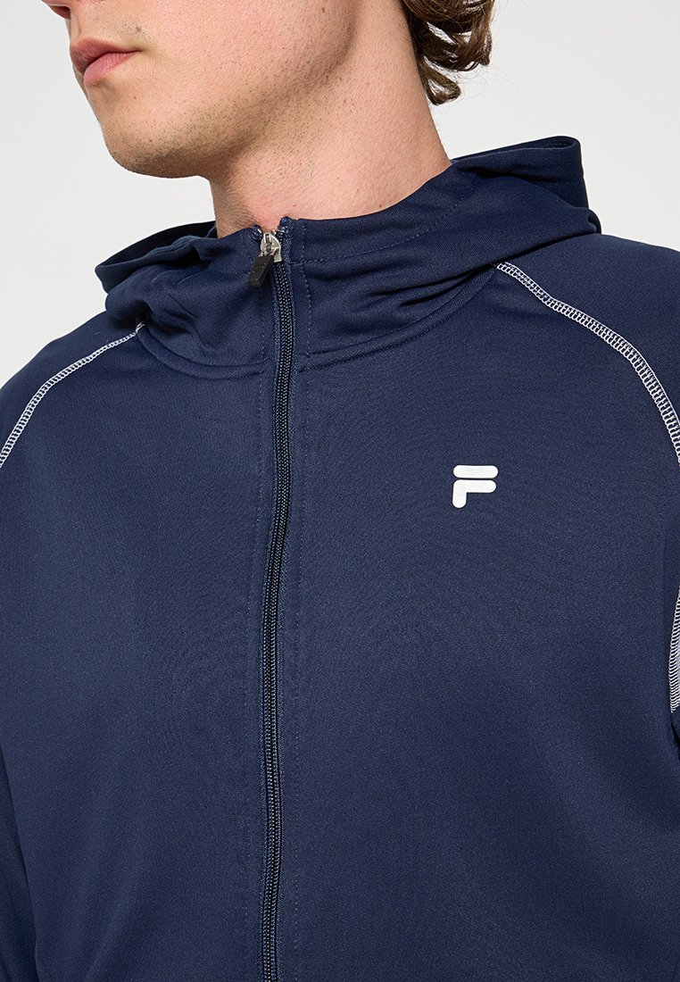 Giacca a zip color navy con cappuccio, caratterizzata da cuciture bianche a contrasto e un piccolo logo sul petto. Realizzata in tessuto liscio e leggero.