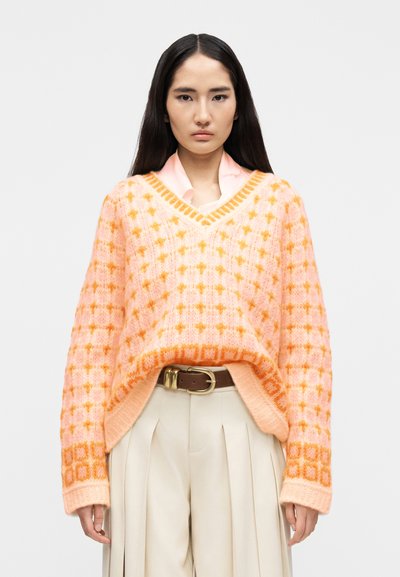 Jeune femme aux longs cheveux noirs portant un pull à col en V à motifs roses et orange par-dessus une chemise rose à col, un pantalon plissé beige et une ceinture marron.