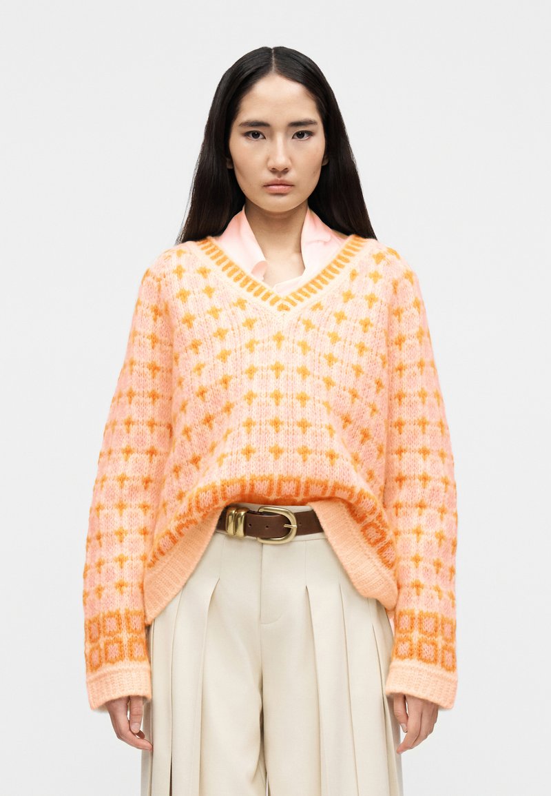 Νεαρή γυναίκα με μακριά σκούρα μαλ λιά, φοράει ροζ και πορτοκαλί πουά πουλόβερ με λαιμόκοψη σε σχήμα V πάνω από ροζ πουκάμισο με γιακά, μπεζ πλισέ παντελόνι και καφέ ζώνη.