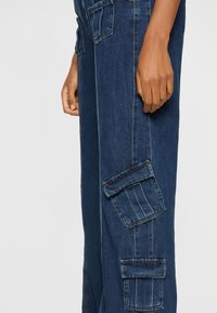 Side view of a person wearing dark blue denim cargo pants with flap pockets, showing their right hand relaxed by their side.