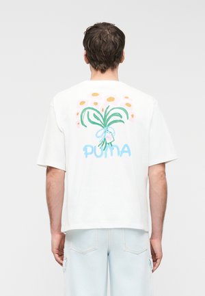 Άνδρας που φοράει λευκό μπλουζάκι με πολύχρωμο floral σχέδιο και κείμενο "PUMA" στην πλάτη, συνδυασμένο με ανοιχτό μπλε παντελόνι.