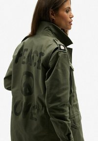 Chaqueta bomber verde oliva con grandes textos en negro "PAZ" y "AMOR" en la parte posterior, con un símbolo de la paz redondo y un parche negro en el hombro.