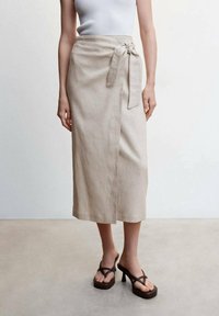 Jupe midi en lin beige avec un design croisé, nouée à la taille. Le tissu a une texture lisse. Associée à des tongs à plateforme marron.