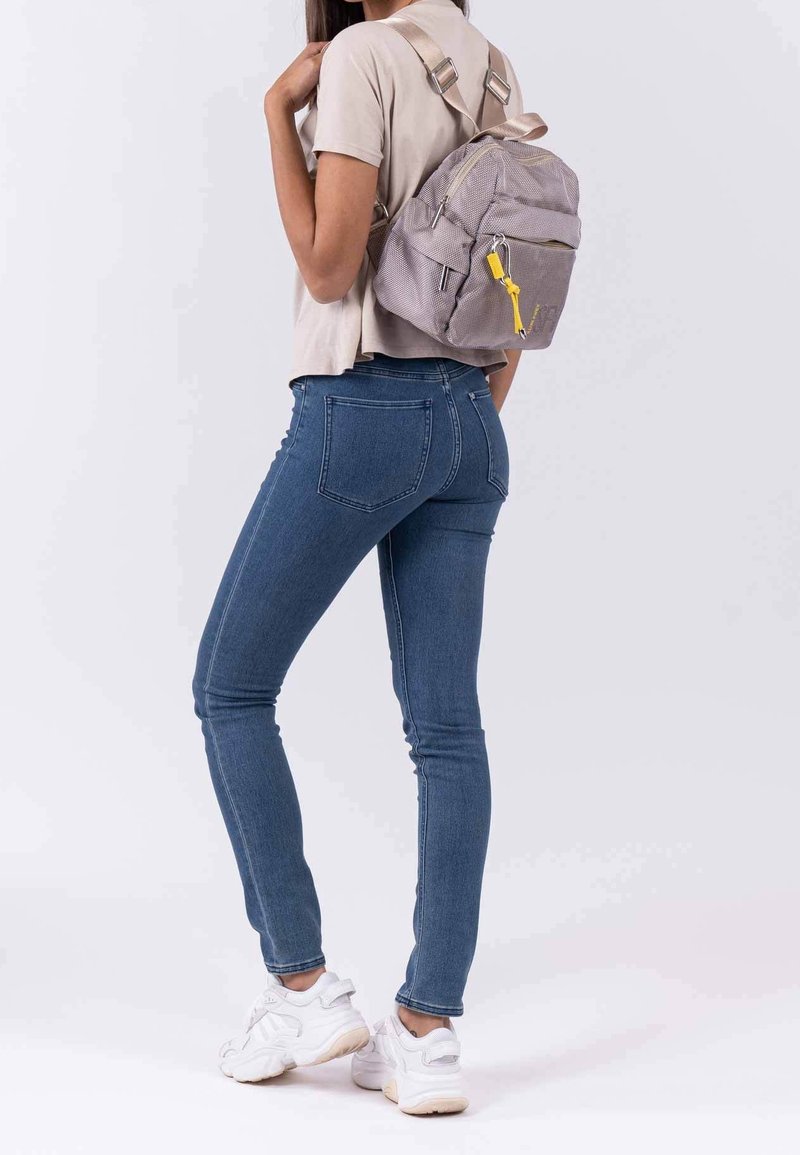 Personne portant un jean bleu, un t-shirt beige, des baskets blanches et un sac à dos gris avec une fermeture éclair jaune, debout devant un fond uni.
