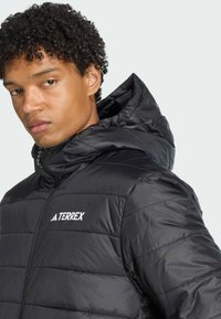 Young person with curly hair wearing a black puffer jacket with a hood, looking directly at the viewer against a light background.