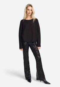 Czarny, oversize sweter dzianinowy z długimi rękawami w połączeniu z czarnymi, cekinowymi spodniami w kształcie dzwonów; całość uzupełniają czarne, szpiczaste szpilki.