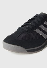 Melns sporta apavu ar mākslīgās zamšāda augšējo daļu un tīklojuma detaļām. Īpašības: pelēkas svītras, teksturēta gumijas ārējā zole un apaļas auklas.