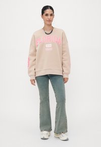 Felpa beige con testo rosa "FLORIDA" e dettagli "MIAMI"; abbinata a jeans chiari a zampa d'elefante e sneakers bianche; orecchini tondi.