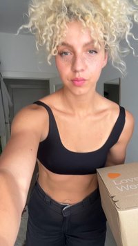 Black sports bra with wide straps, paired with black pants. A cardboard box labeled "Love Well" held in right hand. Light-colored background.