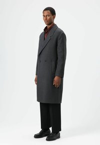Donkergrijze double-breasted wollen jas met zijzakken, kentkraag en gestructureerde schouders, gecombineerd met zwarte broek en schoenen.