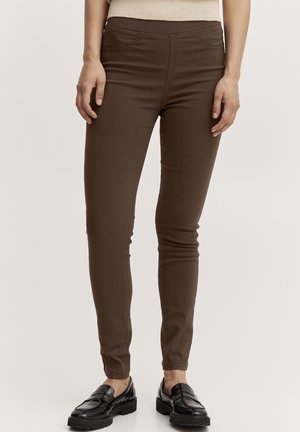 Personne portant un pantalon marron slim avec des poches avant cousues et des mocassins en cuir noir, debout devant un fond uni.