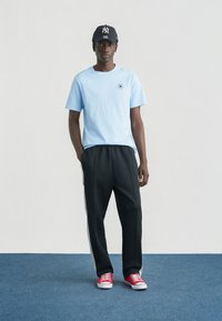 Ανοιχτό μπλε βαμβακερό μπλουζάκι με μικρό λογότυπο, συνδυασμένο με μαύρο παντελόνι φόρμας με λευκές πλευρικές ρίγες και κόκκινα αθλητικά παπούτσια.