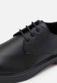 Chaussure habillée noire en cuir à lacets avec une finition lisse, un embout rond et une fermeture à trois œillets sur une semelle en caoutchouc légèrement surélevée.