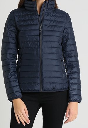 Nainen, joka pukeutuu laivastonsiniseen tikattuun vetoketjulliseen takkiin, jossa on korkea kaulus, ja mustiin housuihin, kädet rentoina sivuilla.