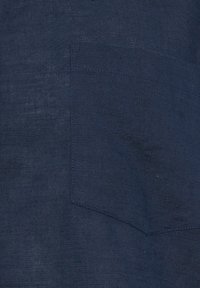 Tissu bleu foncé avec une poche rectangulaire cousue sur le côté gauche, montrant la texture et des détails subtils du tissage.
