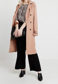 Donna che indossa un trench beige sopra una camicia con stampa zebrate, pantaloni neri a gamba larga, mule nere con tacco, tiene in mano una borsa nera e un bracciale in tartaruga.