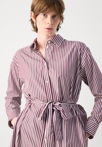 Abito camicia a righe in bordeaux e bianco, con colletto, chiusura a bottoni, maniche lunghe e una vita legata con un dettaglio a fascia.