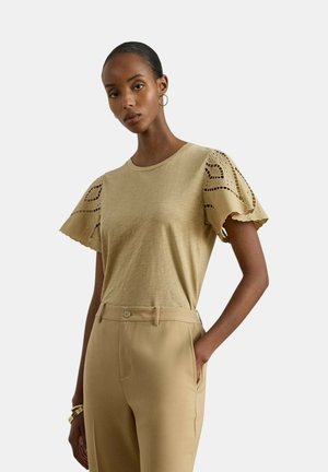 Donna con capelli tirati indietro indossa una camicetta beige con maniche a sbuffo ricamate e pantaloni a vita alta coordinati, in piedi con una mano in tasca.