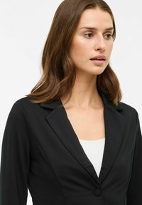 Juodas blazeris su prigludusia forma, įpjautu apykakle, vienu mygtuko užsegimu ir ilgomis rankovėmis. Dėvimas virš balto topuko. Lygus paviršius.