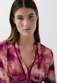 Blůza má kombinaci růžových a oranžových tie-dye vzorů, s límečkem a designem přes hlavu. Tkanina vypadá měkce s mírně splývavou texturou.
