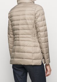 Beige vadderad jacka med vertikalt quiltat mönster, hög krage och sidoöppningar vid fållen. Har en slät, glänsande yta.