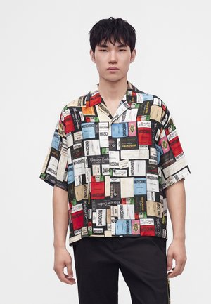 Jeune homme portant une chemise à manches courtes boutonnée recouverte d'imprimés colorés du label Moschino sur un fond uni.