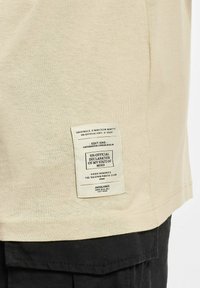 Beige bomull T-skjorte med en strukturert etikett som har design detaljer og tekst, plassert på venstre side nær den nederste sømmen.