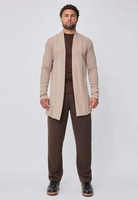 Cardigan long beige clair en tissu doux, à ouverture frontale, porté sur une chemise marron foncé et un pantalon marron assorti, associé à des chaussures noires.