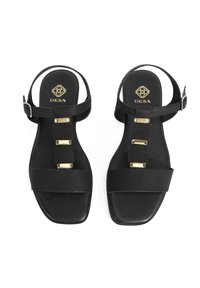 Black sandals with textured straps, gold hardware accents, and a flat sole. Features an open back and square toe design. Brand: DESA.