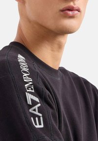 Primo piano della spalla e del collo di una persona che indossa una felpa nera con il testo bianco del marchio "EA7 Emporio" sulla manica.