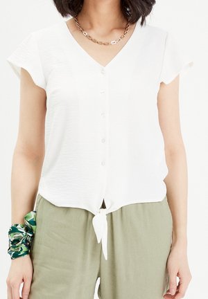 Femme portant un chemisier blanc à manches courtes boutonné, noué à la taille, pantalon vert, collier chaîne en or et chouchou vert à motif au poignet.