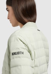Jachetă pufos de culoare verde deschis, cu guler cu nervuri, textură matlasată și textul negru vizibil "KHUJOTM" pe mâneca stângă.