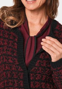 Strickjacke in Schwarz und Burgunderrot mit einem strukturierten Muster. Darunter ist eine kastanienfarbene Bluse mit einem schwarzen Bindeband sichtbar.