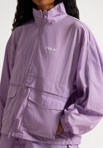 Γυναίκα που φορεί ένα ανοιχτό μωβ μπουφάν με φερμουάρ, με λογότυπο «POLO», τσέπες με καπάκι μπροστά και ελαστικά μανίκια, με το χέρι της να ακουμπά σε μία από τις τσέπες.