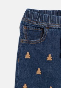 Denimist püksid elastse vöökohaga, millel on laiali triibitud beežkaru tikandid, viie tasku disain ja kontrastne oranž õmblus.