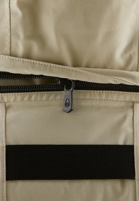 Beige Stofftasche mit einer glatten Textur, ausgestattet mit einem schwarzen Reißverschluss und einem Nylon-Webriemen. Der Reißverschlussgriff hat eine winklige Form mit einem Logodesign.