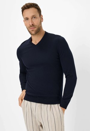 Navy gestrickter Pullover mit V-Ausschnitt und langen Ärmeln, ausgestattet mit gerippten Bündchen und Saum. Getragen mit hellgestreiften Hosen.