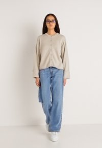 Beige κουμπωτή κάρντιγκαν με χαλαρή εφαρμογή και μακριά μανίκια, συνδυασμένο με ανοιχτό μπλε παντελόνι πλατύτερης γραμμής και λευκά αθλητικά παπούτσια. Λείος υφασμάτινος τύπος.