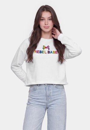 Giovane donna con lunghi capelli castani che indossa una felpa bianca con una scritta colorata "REBEL BABE" e jeans azzurri, posando con la mano vicino al collo.
