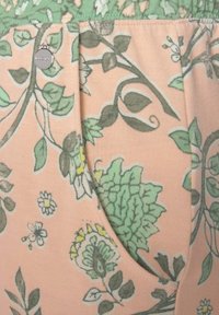 Floral patterned fabric in soft pink featuring green and yellow accents. Includes a pocket and silver logo detail. Smooth texture.