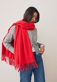 Écharpe rouge aux bords effilochés drapée sur un cardigan gris et un haut blanc, portée avec un jean bleu. Texture douce, contraste de couleurs vives.