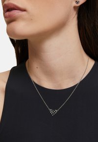 Zilveren ketting met een hartvormige hanger met een open ruitpatroon, gedragen met oorbellen met studs, op een persoon met een gladde halslijn.