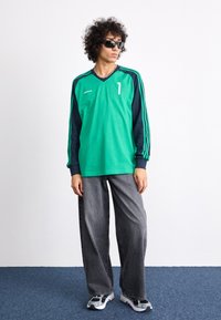 Green and navy long-sleeve sports shirt with a v-neck, featuring a white number "1" and three stripes. Paired with loose grey pants and sneakers.