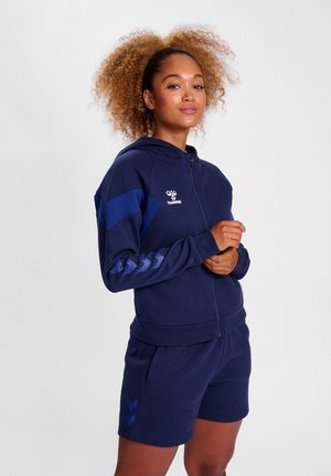 Mørkeblå zip-up hoodie med matchende shorts, lavet af blødt materiale. Har chevronmønstre på ærmerne og shortsene, med logo på brystet.