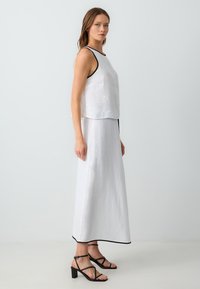 Haut sans manches en lin blanc avec des finitions noires, associé à une jupe maxi en lin blanc. Des sandales à talons blocs noires avec lanières complètent la tenue.