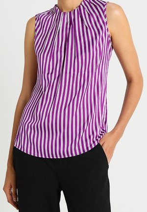 Blouse violette sans manches avec des rayures verticales blanches et des plis au niveau de l'encolure, portée avec un pantalon noir, main dans la poche.
